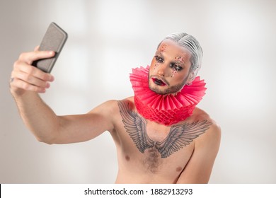 Young Contemporary Actor With Stage Makeup, Tattoo On Chest And Red Paper Jester Collar Around Neck Making Selfie Against White Background