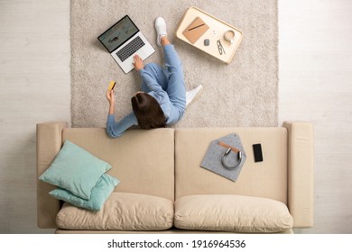 Young Contemporary Brunette Female In Blue Pajamas Resting On The Floor By Couch During Online Shopping At Leisure In Home Environment