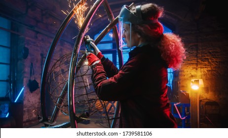 Young Contemporary Female Artist Is Grinding Her Metal Tube Sculpture With A Handheld Power Tool In A Studio Workshop. Empowering Woman Makes Modern Brutal Abstract Artwork Out Of Metal.