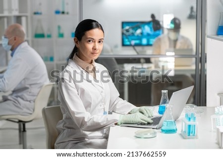 Young contemporary researcher in lab coat and gloves typing in front of laptop and looking at camera while sitting by desk