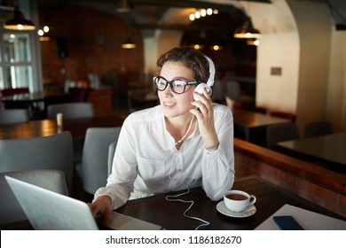 Young Contemporary Woman With Headphones Sitting In Cafe In Front Of Laptop And Watching Webcast By Cup Of Tea