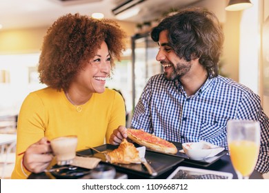 Young Cool Couple Having A Breakfast At Cafe. They Are Drinking Orange Juice And Eating A Croissant And A Toast Bread. They Are Laughing And Having Fun.