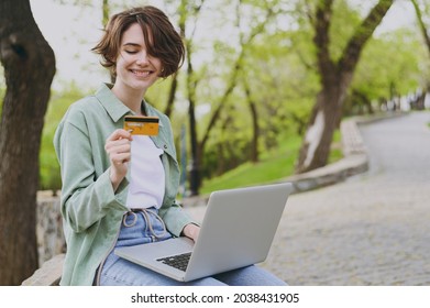 Young Cool Freelancer Woman In Green Jacket Sit On Bench In Spring Park Outdoors Rest Use Laptop Pc Computer Hold Credit Bank Card Do Online Shopping Order Booking Tour. People Urban Lifestyle Concept