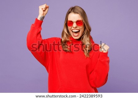 Young cool fun joyful blonde woman she wears red hoody casual clothes sunglasses do winner gesture scream shout yes isolated on plain pastel light purple background studio portrait. Lifestyle concept