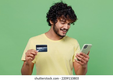 Young Cool Happy Indian Man 20s In Basic Yellow T-shirt Using Mobile Cell Phone Hold Credit Bank Card Doing Online Shopping Order Delivery Booking Tour Isolated On Plain Pastel Light Green Background