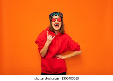 Young And Cool Hipster Girl Having Fun, With Colored Hat And Sunglasses Posing With Good Vibes To Camera On Orange Background