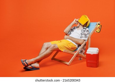 Young Cool Tourist Man Wearing Beach Shirt Hat Lie On Deckchair Near Fridge Read Book Cover Face Sleep Isolated On Plain Orange Background Studio Portrait. Summer Vacation Sea Rest Sun Tan Concept.