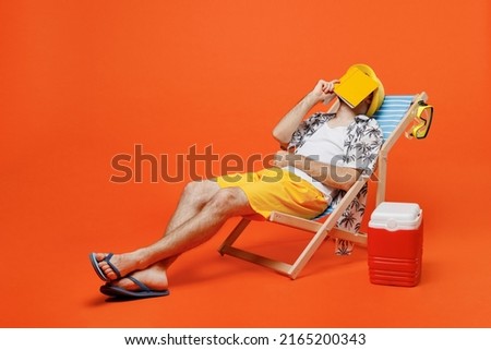 Young cool tourist man wearing beach shirt hat lie on deckchair near fridge read book cover face sleep isolated on plain orange background studio portrait. Summer vacation sea rest sun tan concept.