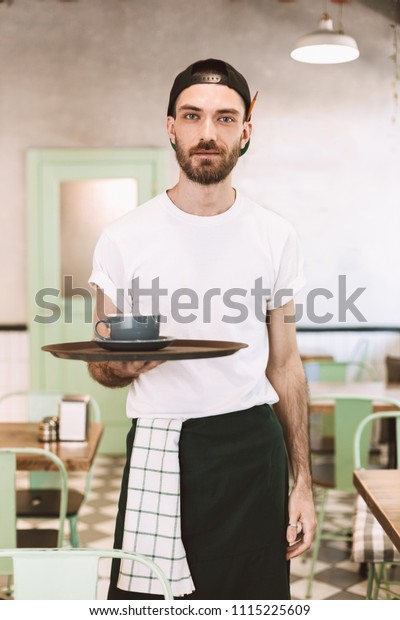 黒い帽子とエプロンをかぶった若い冷たいウェイターが カフェで夢見るようにカメラを見ながら コーヒーを入れたカップとトレイを持つ の写真素材 今すぐ編集