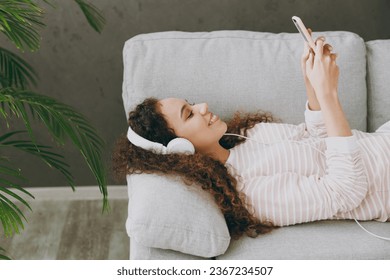 Young cool woman wearing casual clothes lay down on grey sofa couch listen to music in headphones use mobile cell phone stay at home hotel flat rest relax spend free spare time in living room indoor - Shutterstock ID 2367234507