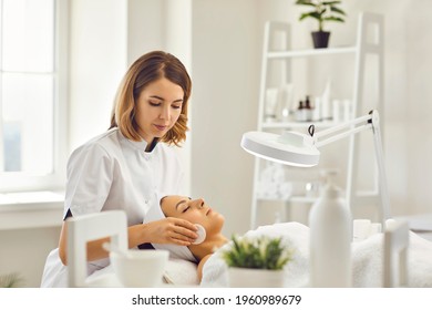 Young cosmetologist or dermatologist cleaning lying womans face with cotton pads in spa salon