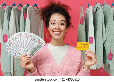 Young Costumer Woman 20s Wear Sweater Stand Near Clothes Rack With Tag Sale In Store Showroom Hold Fan Of Cash Money In Dollar Banknotes Credit Card Isolated On Plain Pink Background Studio Portrait.