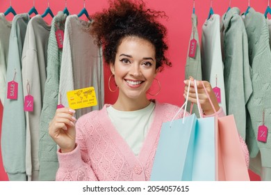 Young Costumer Woman In Sweater Stand Near Clothes Rack With Tag Sale In Store Showroom Hold Package Bags With Purchases After Shopping Credit Card Isolated On Plain Pink Background Studio Portrait