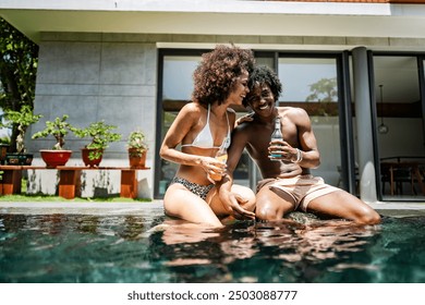 A young couple of African descent enjoying a sunny day at a luxurious poolside, sitting at the edge of the pool with drinks in hand. Modern villa in the background enhances the tropical vacation vibe - Powered by Shutterstock