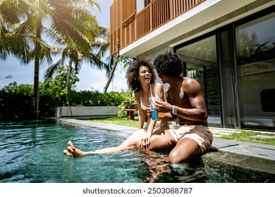 A young couple of African descent enjoying a sunny day at a luxurious poolside, sitting at the edge of the pool with drinks in hand. Modern villa in the background enhances the tropical vacation vibe - Powered by Shutterstock