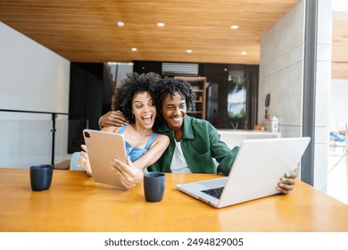 A young couple of African descent in a modern living room, smiling and enjoying their digital devices. A woman holds a tablet, and the man uses a laptop, creating a cheerful and connected atmosphere
 - Powered by Shutterstock