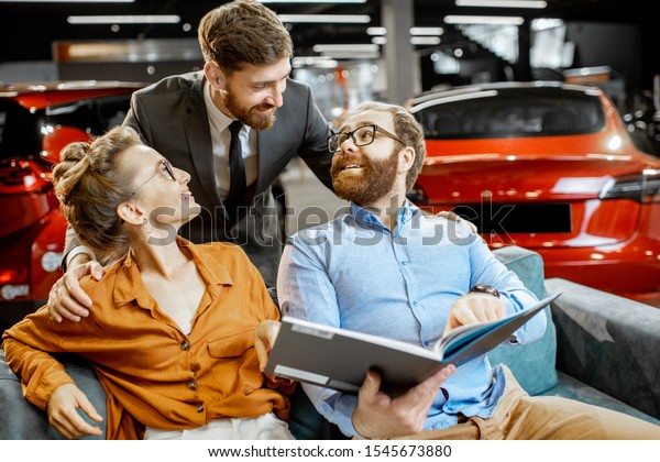 Young couple with annoying salesman choosing\
car finish using catalog, sitting on the comfortable sofa at the\
car dealership