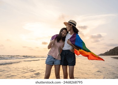 Young couple asian lesbian with pride movement LGBT holding rainbow flag for freedom. Demonstrate rights LGBTQ celebration pride Month lesbian Pride Symbol. Walking on the sand sea beach with sunset