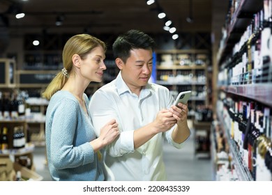 Young Couple, Asian Man And Blonde Woman Choose Wine In Supermarket, Use Phone With App