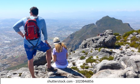 A Young Couple With Backpack And Sun Hat Has Climbed Table Mountain And Looks Down On Cape Town