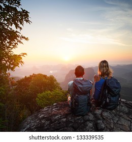 Young couple with backpacks relaxing on top of a mountain and enjoying valley view during sunrise
