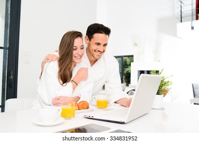 Young Couple In Bathrobe At Home Having Coffee In The Kitchen And Working On Laptop Computer