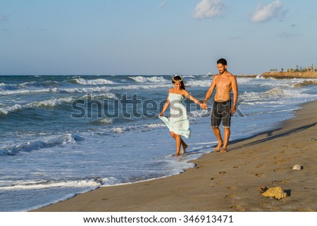 Young couple at the beach on a  hazy summer day at dusk, wearing a turquoise dress and shorts, enjoying  walking barefoot in the ocean water, getting wet, teasing and kissing one another.