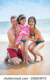 A Young Couple At The Beach Is Posing In Swimsuits With Their Daughter, Mom And Dad Are Drying Their Six Year Old Girl With A Towel, Family Looking At The Camera. 