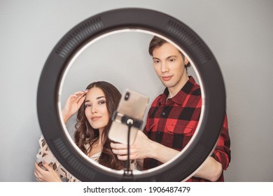 Young Couple Of Beauty Bloggers, Hair Stylist, Hairdresser And Model With Hair Style Shoot On Mobile Phone With Ring Lamp On Grey Wall Background