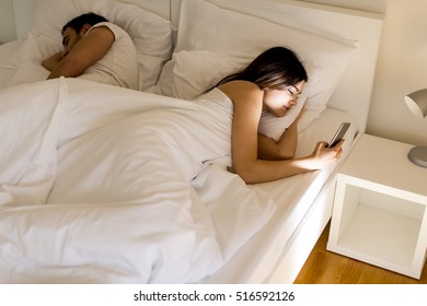 Young Couple In Bed. Man Sleeps While A Young Woman Looking Phone At Night