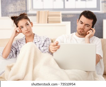 Young Couple In Bed, Man Using Laptop And Mobile, Woman Bored.