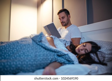 Young Couple In The Bedroom At Night. Man Reading A Book.