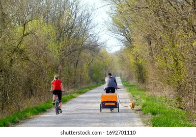 Young Couple Bicycling On Trail In Park On A Nice Spring Day In Midwest; Man Has A Baby Trailer Behind His Bicycle And Small Dog On Leash