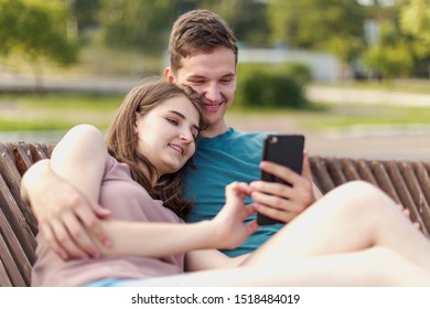 Young couple, a boy and a girl look at the smartphone screen during a summer walk sitting on a bench