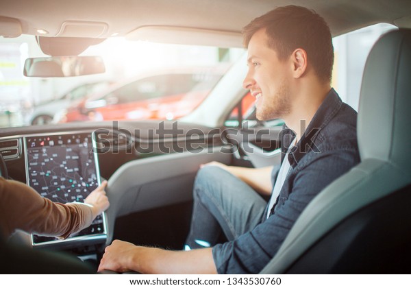 Young couple buying first electric car in the\
showroom. Attractive glad man looking at his girlfriend while she\
chooising a way on electronic dashboard in modern electric hybrid\
vehicle before test