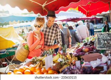 a young couple buying fruits and vegetables in a market on a sunny morning, the young woman carries a basket