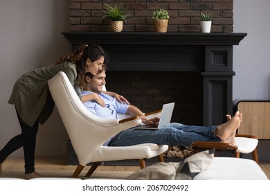 Young couple buying on internet using laptop, wife hugs her husband he sits in comfy armchair looking at computer, discussing purchase, make order, enjoy e-shopping, plan vacation, choose tour concept - Powered by Shutterstock