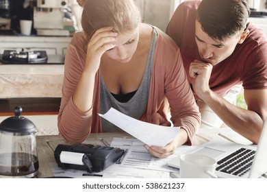 Young couple calculating their domestic budget together in kitchen, trying to save money for buying new car, having stressed and frustrated looks. Unhappy woman showing unpaid bill to her husband