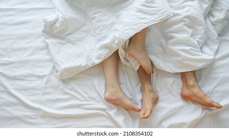 Young Couple Caresses Each Other After Waking Up On Soft Bed On White Sheets Sticking Bare Legs Out Of Blanket In Light Bedroom At Home, Upper View.