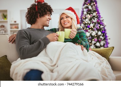 Young Couple Celebrating Christmas At Home, Relaxing With Cups Of Coffee Covered With Blanket On A Sofa In An Apartment.