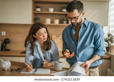 Young couple comparing documents and using a smart phone application to manage their household finances, calculating expenses and making a budget while saving money with a piggy bank