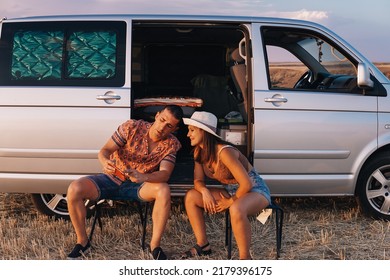 Young Couple Consisting Of Boy And Girl Sitting In Camp Chairs Outside Their Camper Van. The Man Is Looking And Pointing At His Cell Phone With The Intention Of Explaining Something To The Woman.