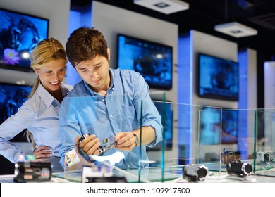 Young Couple In Consumer Electronics Store Looking At Latest Laptop, Television And Photo Camera