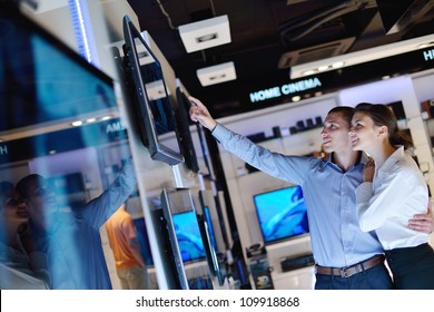 Young Couple In Consumer Electronics Store Looking At Latest Laptop, Television And Photo Camera To Buy