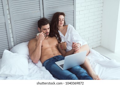 young couple with cups of coffee using laptop while resting on bed at home