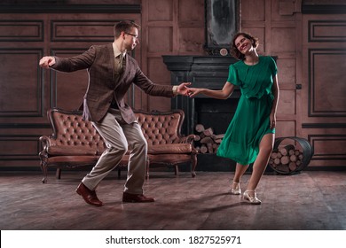 a young couple dancing swing in a retro hall in front of a fireplace and a leathern sofa; the woman wears a green dress