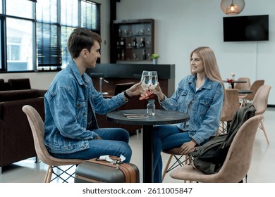 Young couple in denim jackets clinking water glasses at a stylish lounge table, celebrating or enjoying a relaxed travel moment together indoors