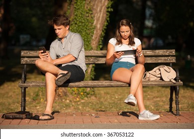 Young couple distracted with communication technology tablet and smart phone sitting on a bench socializing over the web not knowing for each other