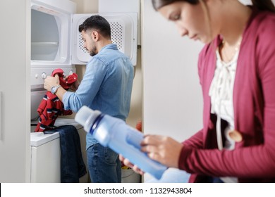 Young Couple Doing Housework And Chores. Man Putting Clothes Inside Washing Machine And Girlfriend Sitting On Kitchen Counter, Reading Label On Cleansing Box.