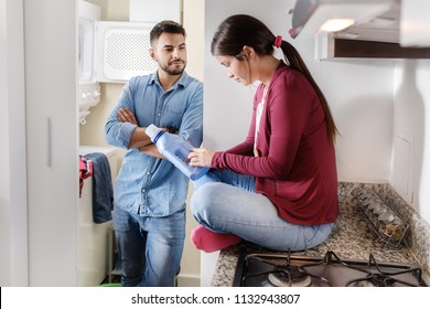 Young Couple Doing Housework And Chores. Man Putting Clothes Inside Washing Machine And Girlfriend Sitting On Kitchen Counter, Reading Label On Cleansing Box.